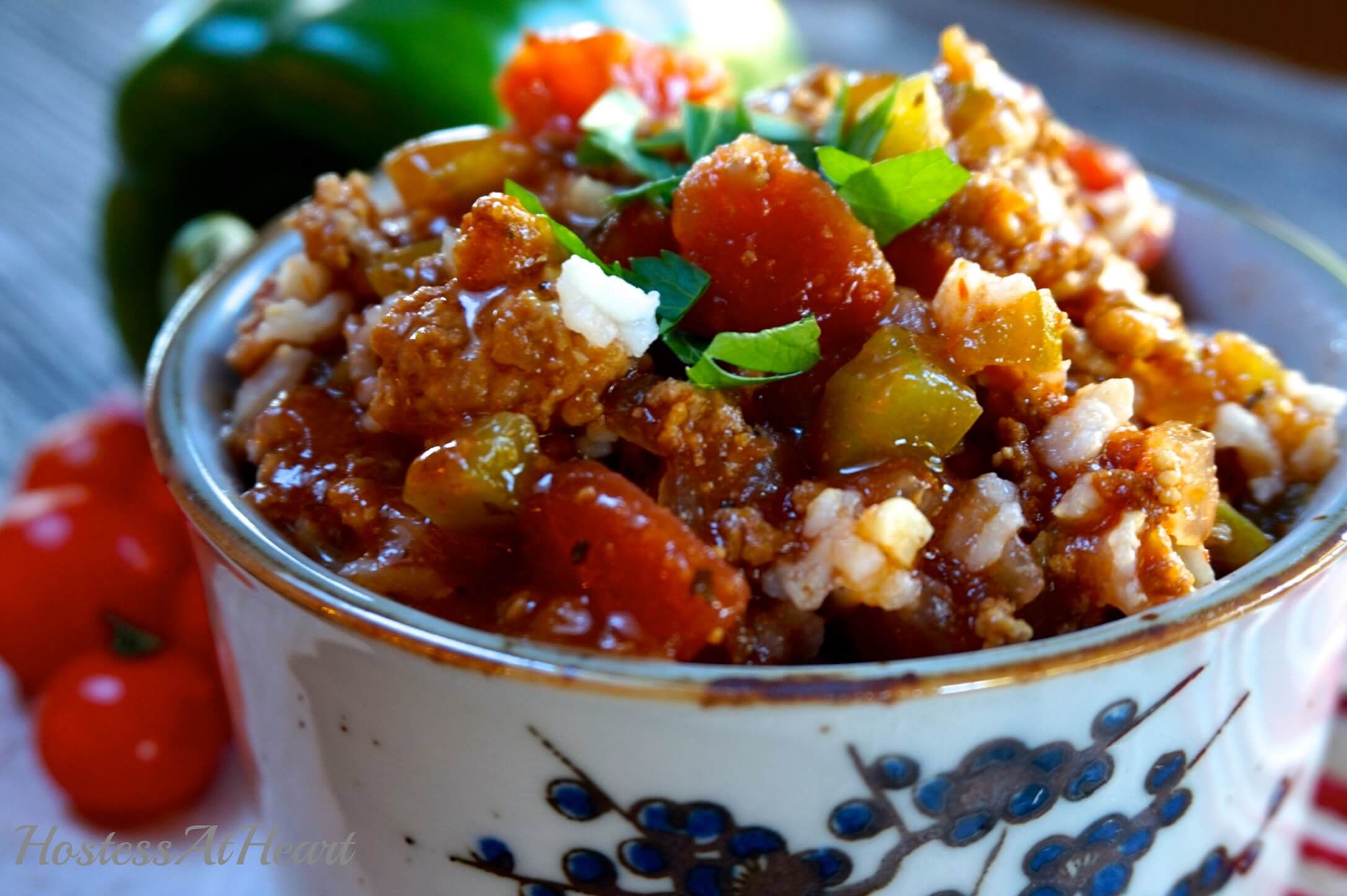 Sideview of a soup bowl with a blue design on the front is heaped with Stuffed Pepper Sour that's full of sausage, peppers, tomatoes, and garnished with fresh parsley. Fresh peppers and tomatoes sit in the background.