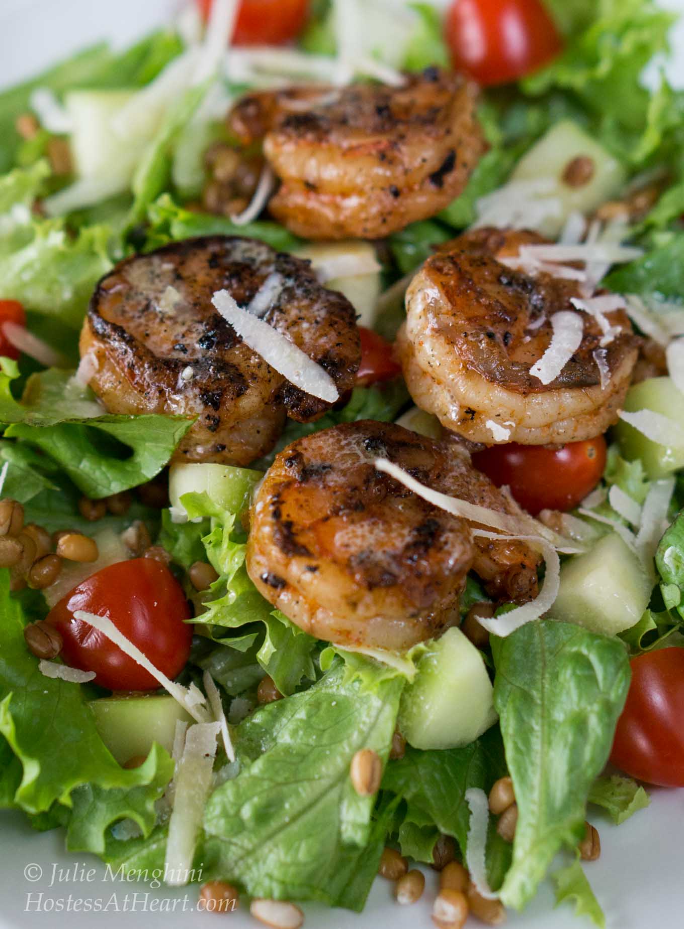 Close-up view of a white plate filled with a green salad, cucumbers, and tomatoes then topped with grilled shrimp and garnished with cheese. 