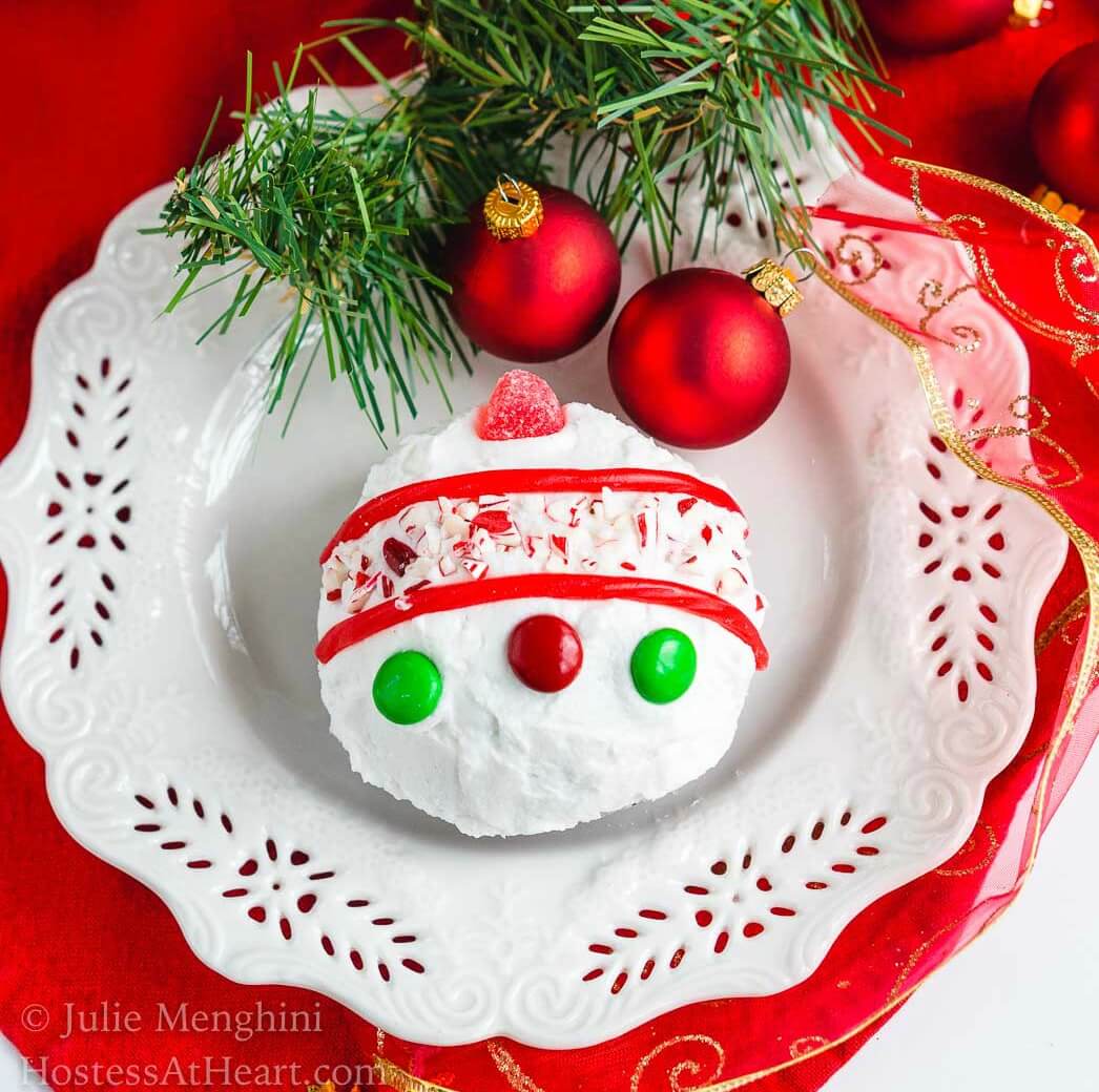 A cake ball frosted in white frosting decorated with candy pieces to resemble a Christmas bulb on a white snowflake plate. A sprig of greenery and two red Christmas bulbs are at the top of the plate.