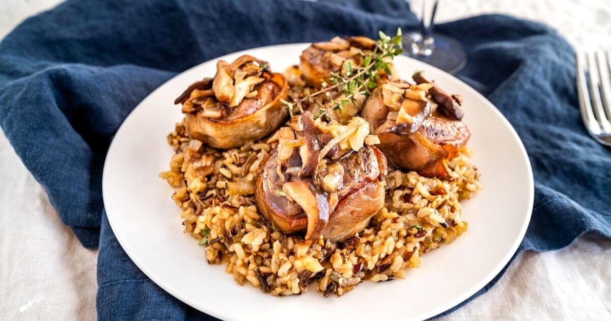 White plate with wild rice in the center topped with 4 bacon-wrapped lamb medallions covered in a wine sauce with mushrooms. A sprig of thyme garnishes the dish.