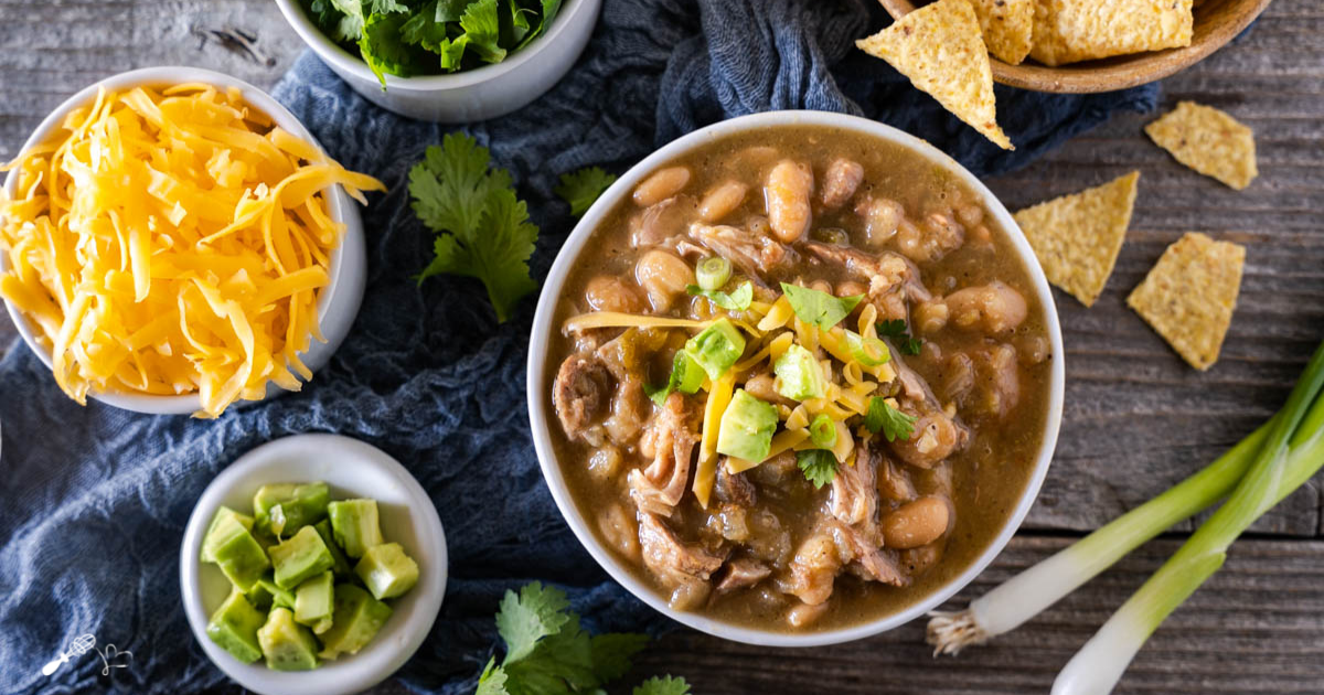 Top down view of a bowl of pork chili topped with avocado and cilantro with bowls of shredded cheese, cilantro, green onions, and tortilla chips off to the side.