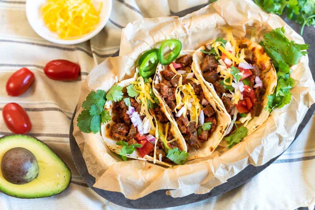 Side angle of three beef brisket tacos in a basket surrounded by avocado and tomato.