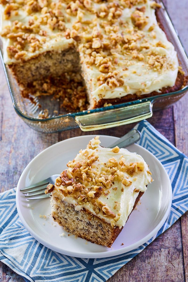 Side close-up view of a single slice of frosted banana cake garnished with nuts. Hostess At Heart